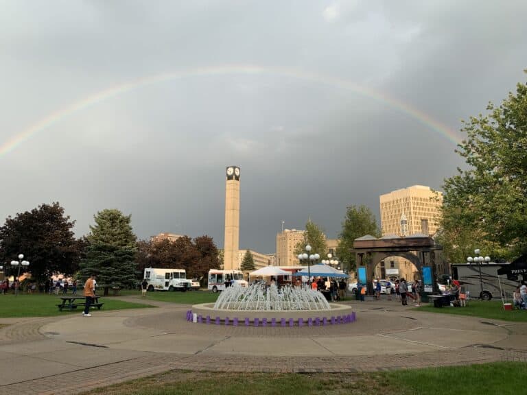 City park with rainbow over fountain and clock tower.