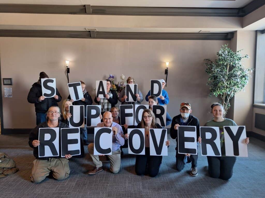 Group holding 'Stand Up for Recovery' signs indoors.