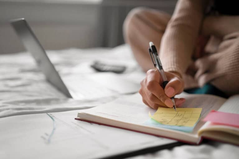Person writing notes on a desk with laptop