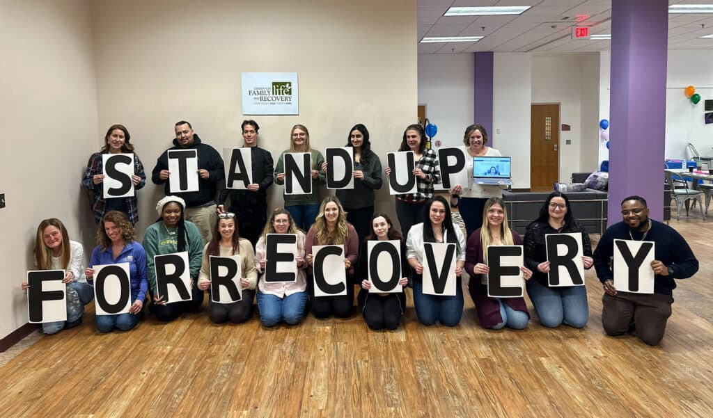 Group holds 'Stand Up for Recovery' signs indoors.